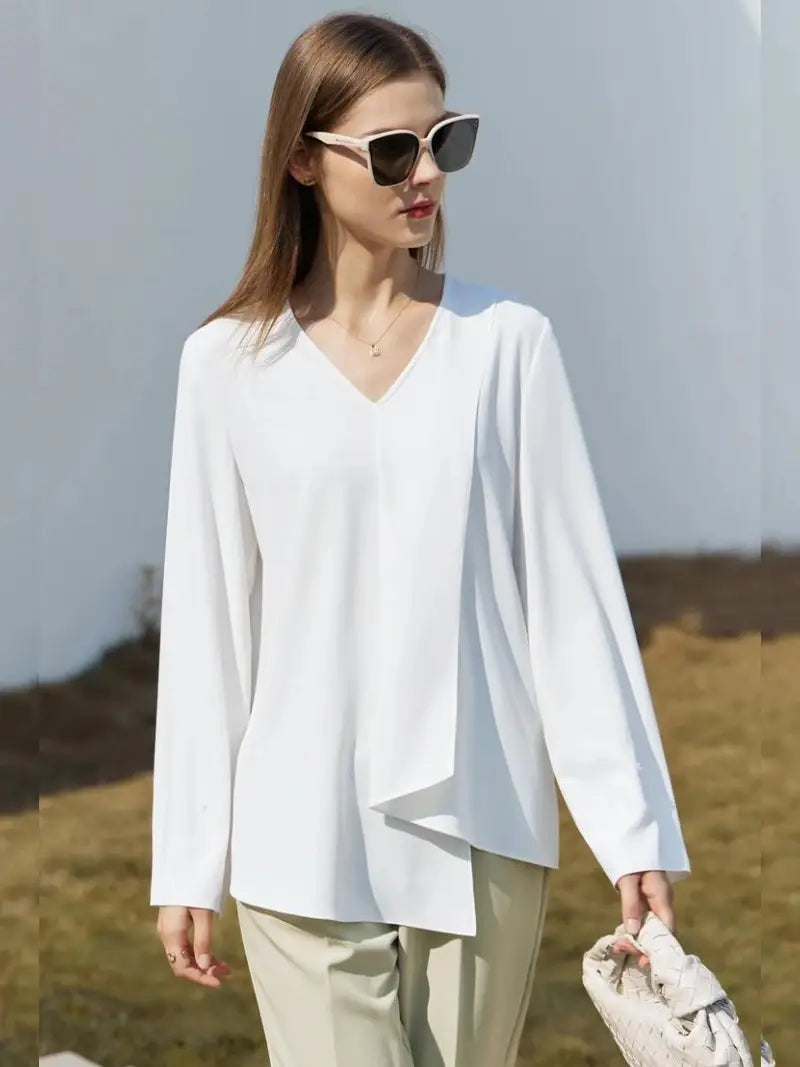 Une femme portant des lunettes de soleil porte un élégant chemisier blanc Vintage à Col en V et un pantalon crème, tenant un sac texturé, debout sur un fond naturel doux.