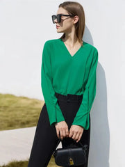 Une femme vêtue d'un élégant BLOUSES VINTAGE À COL EN V vert et d'un pantalon noir se tient avec confiance, tenant un petit sac à main noir, avec des lunettes de soleil. Elle est appuyée contre un mur blanc, à l'extérieur