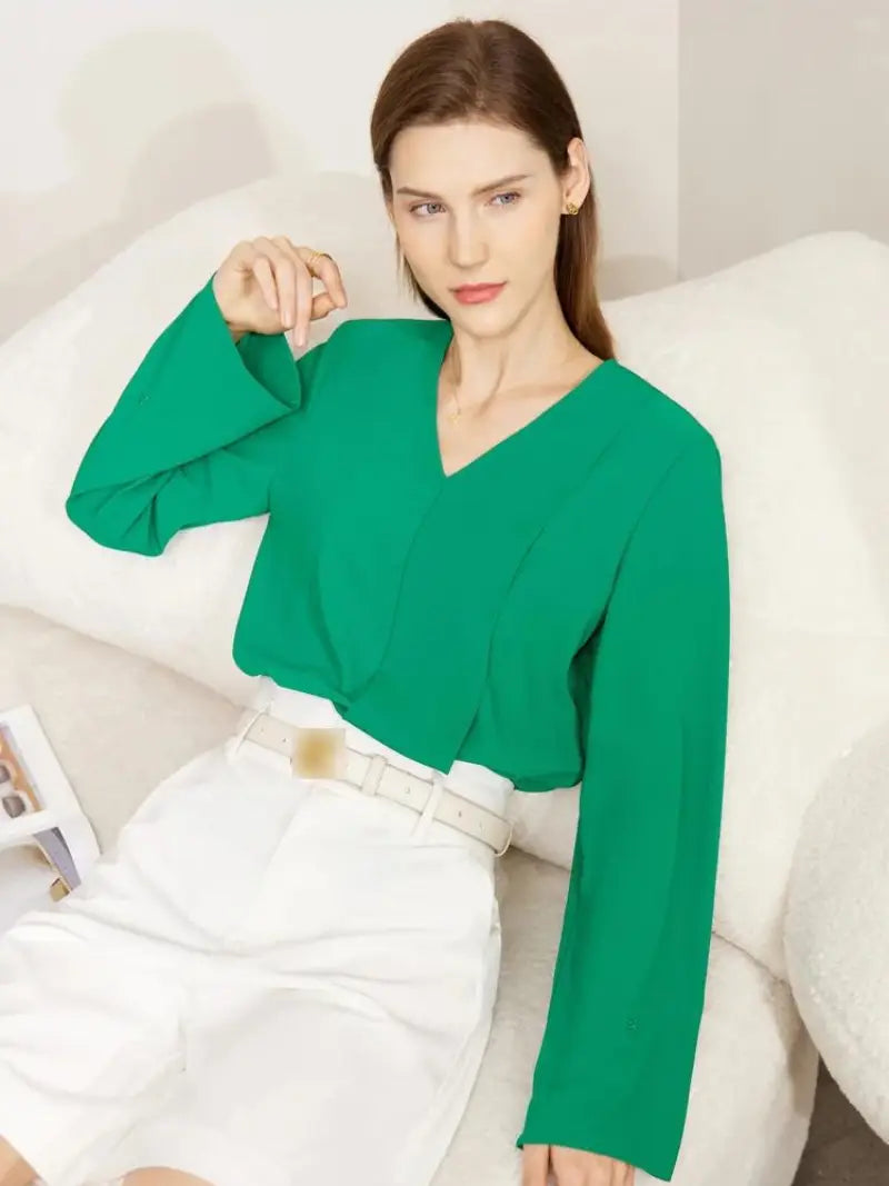 Une femme aux cheveux bruns raides, vêtue d'un chemisier vert vif Vintage à col en V et d'une jupe blanche, assise élégamment sur un fond beige doux. Son expression est calme et posée.