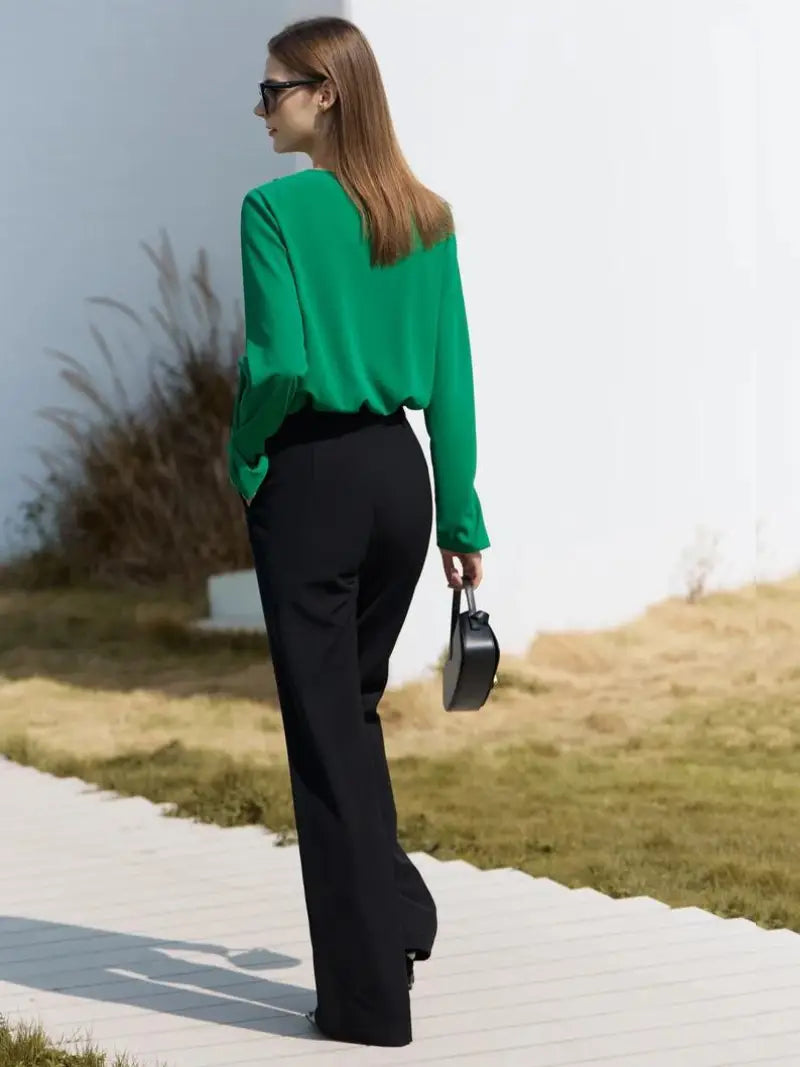 Une femme vêtue d'un BLOUSES VINTAGE À COL EN V vert vif et d'un pantalon noir marche le long d'un sentier blanc, tenant un petit sac à main noir. Elle porte des lunettes de soleil et ses cheveux sont raides et ressemblent à
