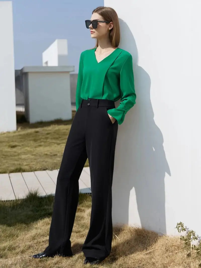 Une femme élégante se tient en toute confiance à l'extérieur, à côté d'un mur blanc, vêtue d'un chemisier vert Vintage à Col en V de l'AMII Printemps 2023, d'un pantalon noir et de lunettes de soleil.