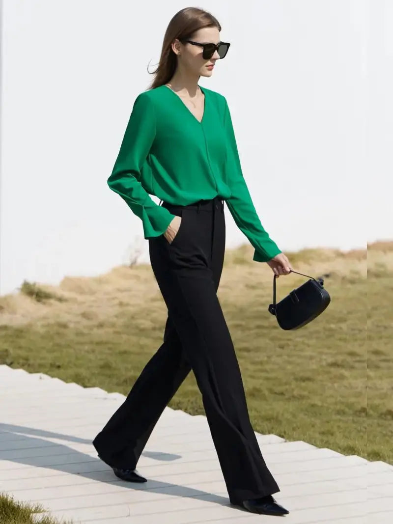 Une femme élégante portant des lunettes de soleil porte un chemisier vert vif Vintage Col en V, un pantalon noir et tient un petit sac à main noir, marchant en toute confiance sur un sentier blanc avec un fond herbeux.