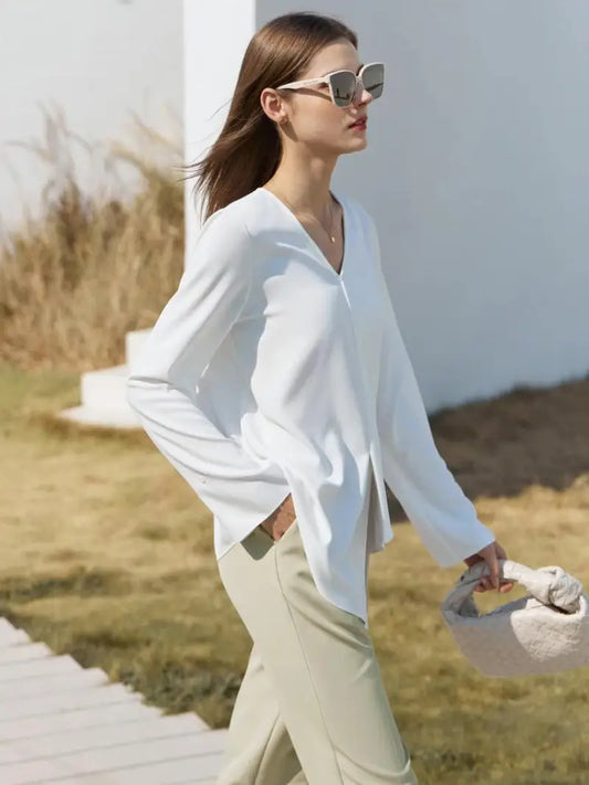 Une femme portant des lunettes de soleil élégantes, un BLOUSES VINTAGE À COL EN V blanc et un pantalon beige marche en toute confiance à l'extérieur, portant un sac texturé, avec une structure blanche et un champ herbeux en arrière-plan.