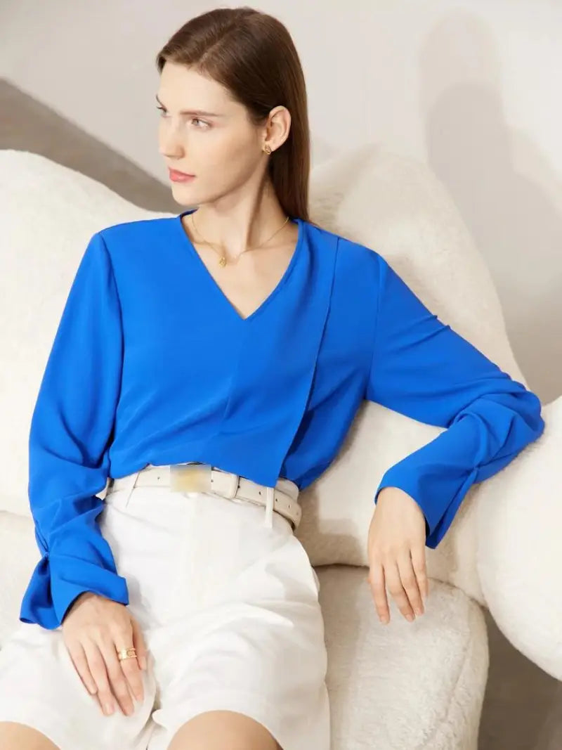 Une femme vêtue d'un chemisier bleu vif Vintage à col en V et d'une jupe blanche est assise élégamment sur un canapé de couleur crème, regardant sur le côté avec une expression sereine. Elle porte