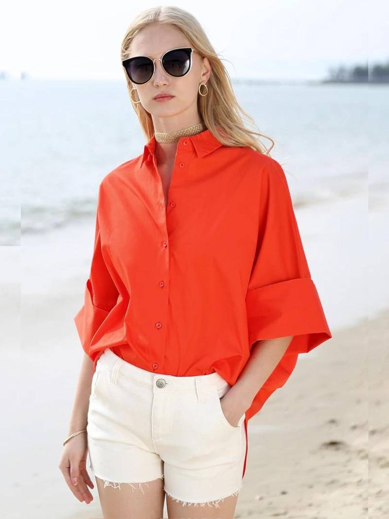 Une femme portant un Vanissy CHEMISIER DE MANCHES 3 QUARTS orange vif en 100 % coton et un short en jean blanc se tient sur une plage, accessoirisée de grandes lunettes de soleil noires.