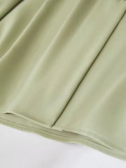 Une jeune femme porte un chemisier Vanissy décontracté en mousseline de soie vert clair avec un décolleté en V et des manches longues, associé à un pantalon blanc et tenant une veste texturée beige.