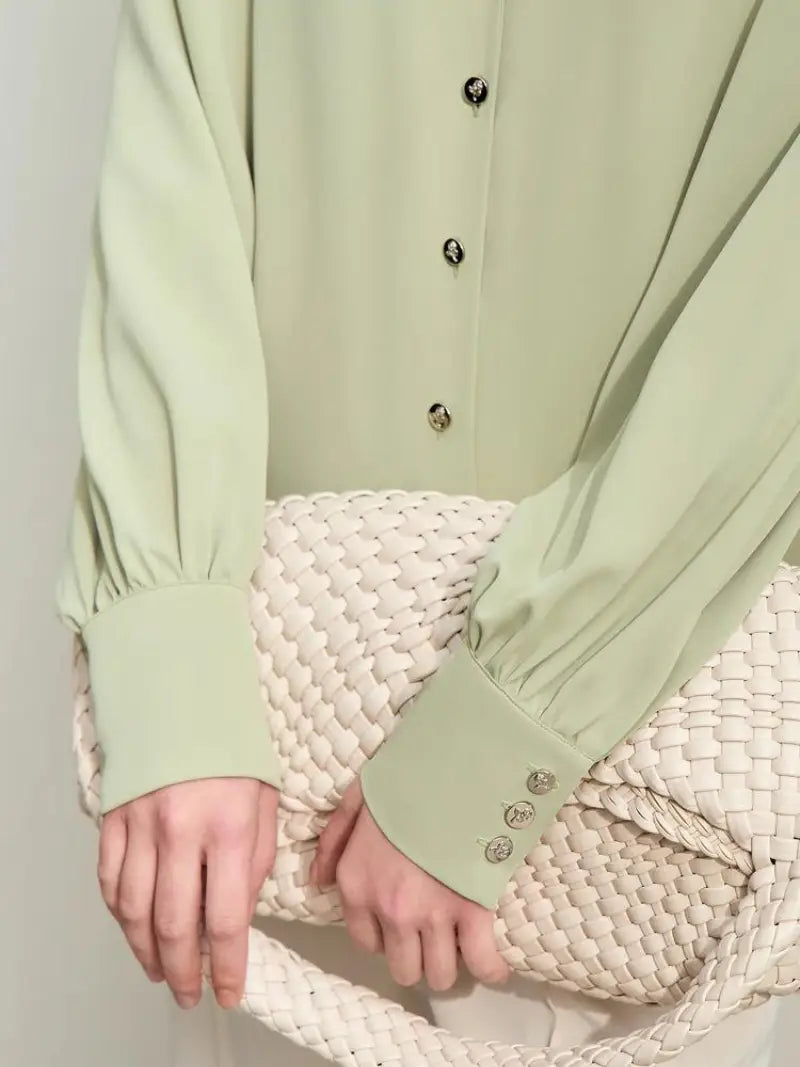 Une jeune femme porte un chemisier Vanissy décontracté en mousseline de soie vert clair avec un décolleté en V et des manches longues, associé à un pantalon blanc et tenant une veste texturée beige.