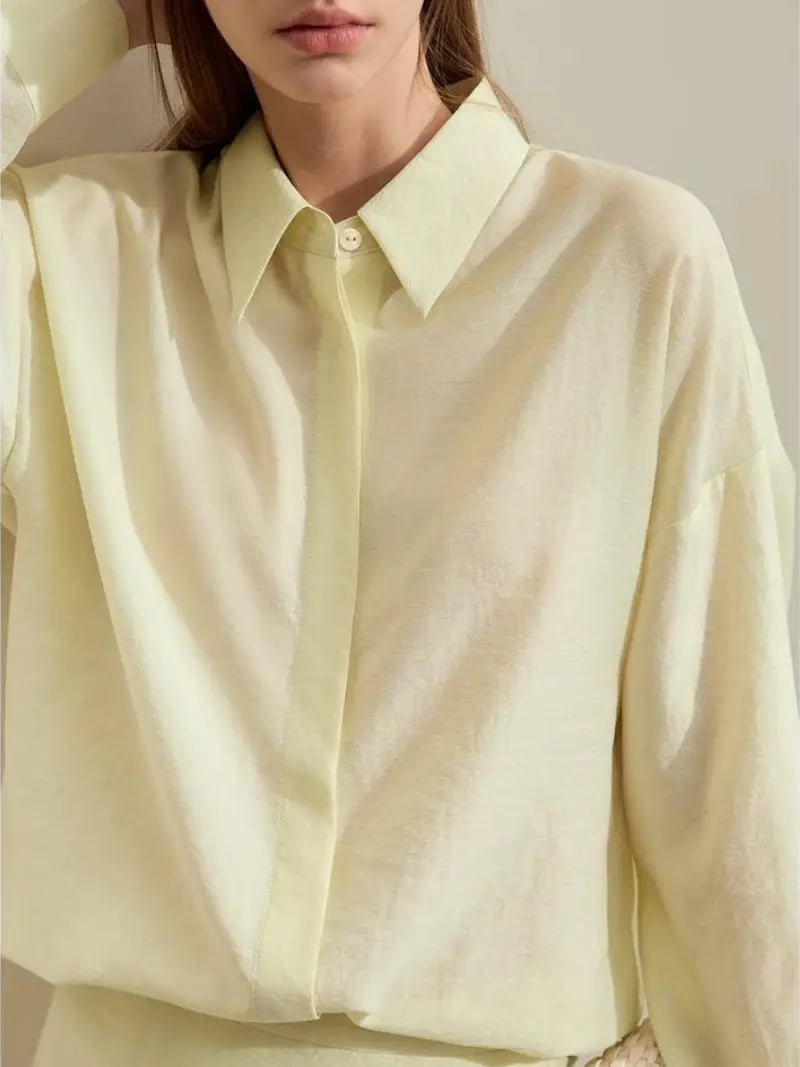 Une femme se tient sur un fond neutre, vêtue d'une chemise et d'une jupe jaune pâle avec une grande ceinture de la collection ENSEMBLE ROBES ET CHEMISIERS Printemps 2024. Elle porte un sac à main texturé et regarde droit devant elle.