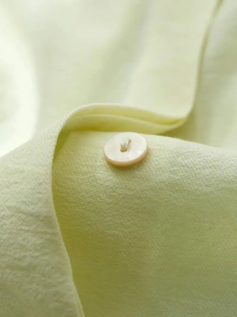 Gros plan d'un petit bouton rond blanc cassé fixé sur un ENSEMBLE ROBES ET CHEMISIERS de couleur et de texture similaires, soulignant les détails subtils du tissu et des coutures.